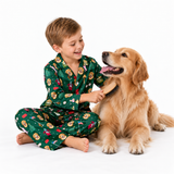 A young boy wearing green Christmas-themed pajamas sits on the floor and brushes a golden retriever dog. The boy's pajamas feature a festive pattern of Santa Claus, reindeer, snow globes, and holiday lights.