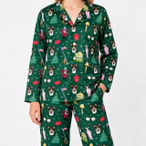 A person wearing a matching long-sleeve pajama set with a festive Christmas print on a dark green background. The pajamas feature an all-over pattern of dog faces, Santa Claus, Christmas trees, bells, houses, and holiday decorations.
