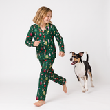 A young girl wearing a green long-sleeve pajama set with a festive Christmas print is walking barefoot next to a black and white dog on a white studio background.