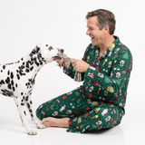 A man wearing a green Christmas-themed pajama set with cow print sits on a white studio floor playing tug-of-war with a Dalmatian dog.