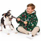 A child wearing a green holiday-themed fleece hoodie blanket sits on the floor and plays tug-of-war with a black and white dog on a white studio background.