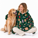 A woman sitting cross-legged on a white studio floor wearing a green hooded fleece pajama set with a colorful dog-themed print, while petting a golden retriever dog.
