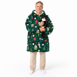 A smiling man wearing a green Christmas-themed hooded robe with a cat print pattern, holding a black and white cat. He is wearing beige pants and white socks against a solid white background.