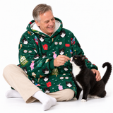 A man wearing a green cat-themed fleece hoodie sits on the floor, smiling as he feeds a black and white cat. The hoodie features a playful print of various cats, a cat bed, and Christmas elements.