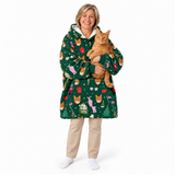 A smiling woman wearing a green Christmas-themed cat-zoned hoodie blanket robe while holding an orange tabby cat, standing against a plain white studio background.