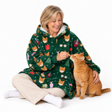 A woman sitting on a white studio floor wearing a green holiday-themed hooded sweatshirt with a cat print pattern, interacting with an orange tabby cat. The sweatshirt features illustrations of cats, Christmas trees, and festive decorations.
