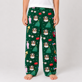 A person wearing dark green holiday lounge pants with an all-over print of Australian Shepherd dogs, Christmas trees, ornaments, and snowflakes, standing barefoot against a light gray studio background.