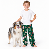 A young boy wearing a white t-shirt and green Christmas-themed pajama pants with a dog print, standing next to an Australian Shepherd dog on a white studio background.