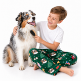 A smiling young boy in a white t-shirt and green Christmas-themed pajama pants brushes a fluffy Australian Shepherd dog on a clean white background.