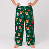 A person wearing dark green holiday-themed lounge pants with an all-over print of orange foxes, Christmas trees, Santa Claus, and snowflakes, paired with a white top against a light gray studio background.