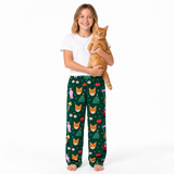 A young girl stands barefoot against a white studio background, holding an orange tabby cat. She is wearing a white short-sleeve T-shirt and dark green pajama pants covered in a festive Christmas pattern featuring orange cat faces, Christmas trees, gifts, and ornaments.