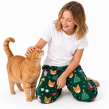 A young girl kneeling on a white studio floor, smiling and petting an orange tabby cat. She is wearing a plain white t-shirt and dark green pajama pants with a festive cat-and-tree pattern.