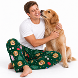 A man wearing green Christmas-themed dog pajama pants sits on the floor hugging a golden retriever dog against a white studio background.
