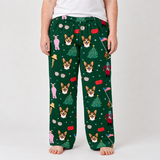 A person wearing dark green Christmas-themed pajama pants with a repeating pattern of corgi dogs, Christmas trees, Santa Claus, and snowflakes, paired with a white t-shirt against a light gray studio background.