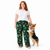 A woman wearing dark green Christmas-themed pajama pants with a corgi dog print stands next to a corgi dog on a white studio background. The pants feature a festive pattern of corgis, Christmas trees, and holiday figures.