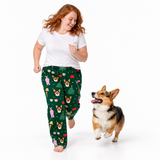 A woman with red hair and a corgi dog running on a white studio background. The woman is wearing a white t-shirt and dark green pajama pants with a festive dog-themed pattern, while the corgi runs happily beside her.