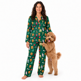A woman wearing a green long-sleeve pajama set with a colorful holiday-themed print, standing barefoot next to a fluffy golden-brown dog on a plain white background.