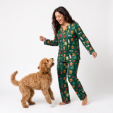 A woman wearing a green long-sleeve pajama set with a holiday gingerbread-themed print is standing barefoot on a white studio background, smiling and reaching toward a fluffy golden-brown dog.