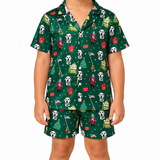 A child wearing a matching short-sleeve button-up pajama shirt and shorts set in dark green with a festive holiday print featuring Dalmatian dogs, Santa Claus, Christmas trees, and houses.