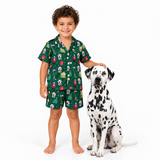 A young boy wearing a green short-sleeve pajama set with a dog print stands next to a Dalmatian dog on a white studio background.