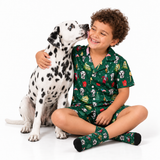 A smiling young boy wearing a green Dalmatian-themed pajama set sits cross-legged on a white studio floor next to a Dalmatian dog, with the dog licking his face.