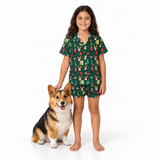 A young girl stands barefoot next to a corgi on a white studio background, wearing a dark green short-sleeve pajama set with a festive Christmas print featuring Santa, elves, gifts, and snowflakes.