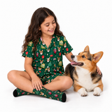 A young girl with long dark hair sits cross-legged on a white studio floor wearing a green Christmas-themed pajama set with a corgi print, next to a corgi dog.