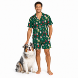 A man wearing a matching green short-sleeve button-up shirt and shorts pajama set with a cartoon dog print, standing next to a fluffy Australian Shepherd dog on a solid white background.