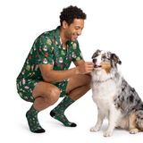 A man wearing a matching green short-sleeve shirt and shorts pajama set with a dog print, crouching down to pet an Australian Shepherd dog on a white studio background.