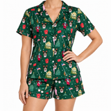 A woman modeling a matching short-sleeve button-front pajama top and shorts set in dark green with a festive holiday print featuring Santa Claus, Christmas trees, houses, and snowflakes.