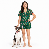 A woman stands barefoot on a white studio background wearing a green short-sleeve pajama set with a Christmas-themed print, next to a black, white, and tan dog.