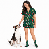A woman wearing a green Christmas-themed pajama set with a dog sitting beside her on a white studio background. The pajama set includes a short-sleeve top and matching shorts, both printed with holiday illustrations such as Christmas trees, ornaments, and Santa, and she is also wearing matching socks.