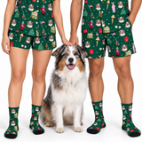 A man and a woman modeling matching green short-sleeve pajama sets and coordinating socks with a dog sitting between them. The pajamas and socks feature a festive Christmas-themed print with dogs, Christmas trees, ornaments, and holiday figures on a white background.