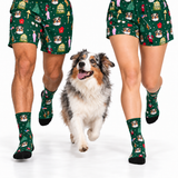 A dog walking between two people wearing matching green Christmas-themed socks and shorts with a dog print, on a white studio background.