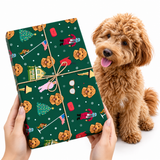 A person's hands hold a gift box wrapped in dark green holiday-themed wrapping paper with a pattern of cookies, Christmas trees, and nutcracker soldiers, tied with brown string. A fluffy golden-brown dog sits to the right on a clean white background.