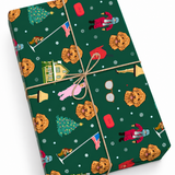 A wrapped rectangular gift box shown at an angle on a white background. The box is covered in dark green holiday-themed wrapping paper with a repeating print of golden doodog faces, Christmas trees, snowflakes, flags, glasses, and a pink figure, tied with natural twine.