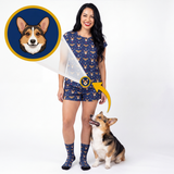 A woman modeling a matching navy blue pajama set with a corgi print, standing next to a corgi dog on a white studio background. A circular inset shows a corgi face, with a yellow arrow pointing to the printed design on the pajamas and socks.