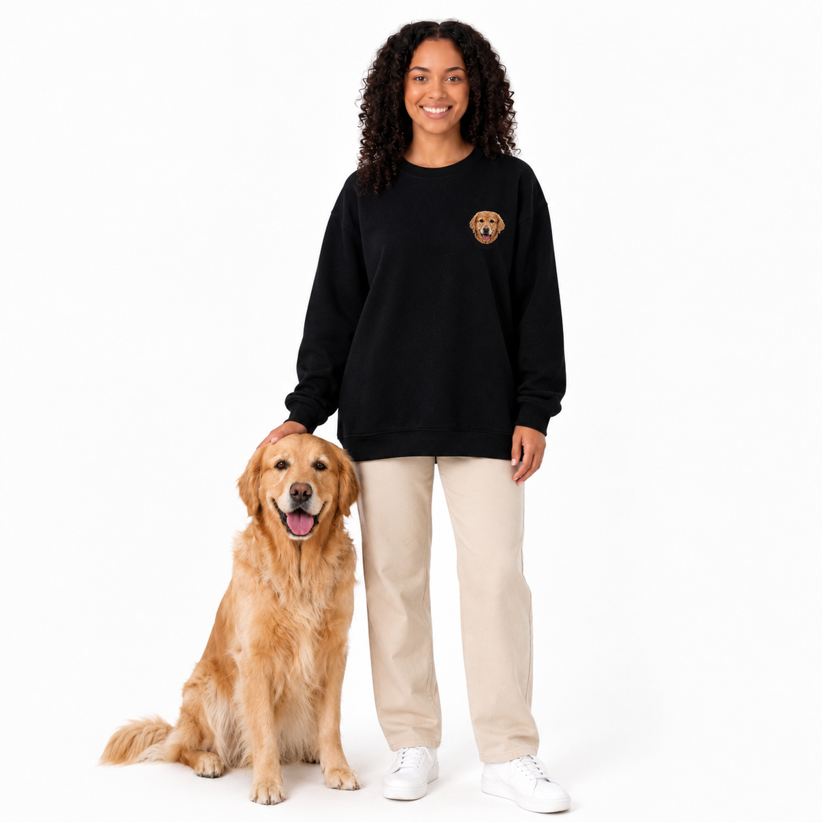 A woman stands next to a golden retriever dog on a white studio background, wearing a black crewneck sweatshirt with a small dog-face patch on the left chest, beige pants, and white sneakers.