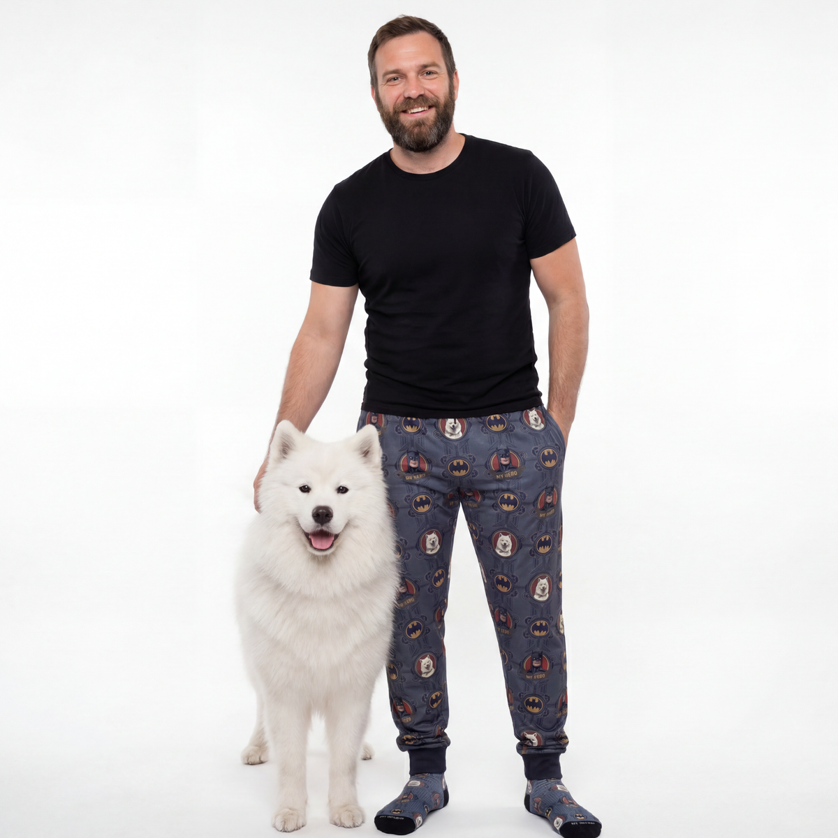 Man wearing a black t-shirt and patterned pants standing next to a white dog on a white background