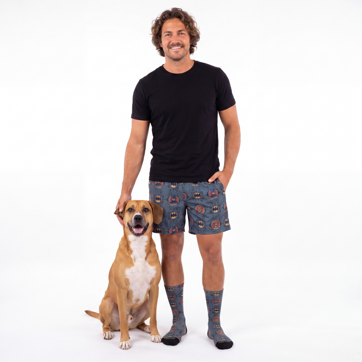 Man wearing black t-shirt and patterned shorts standing next to a dog on a white background
