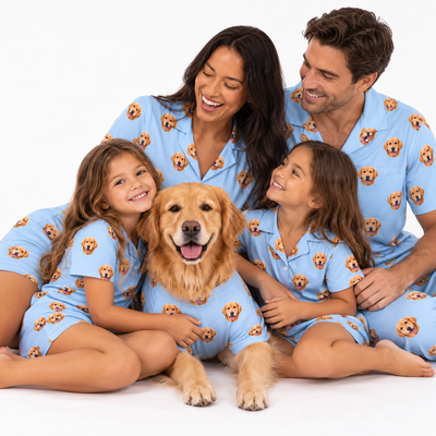 A family of four and their golden retriever dog wearing matching light blue pajamas printed with a repeating dog face pattern, posed together on a white studio background.
