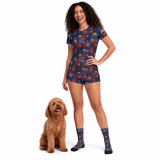 A woman wearing a matching Supergirl-themed short-sleeve fitted shirt set and socks with dog print, standing next to a small brown poodle on a white background.