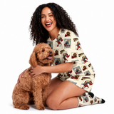 A woman kneeling on a white studio background, smiling and hugging a small brown dog. She is wearing a cream pajama set with a repeating comic-book style supergirl print.
