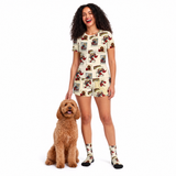 A woman wearing a short-sleeve cream-colored shirt and short set with a comic book vintage-style print, standing next to a brown poodle on a solid white background.