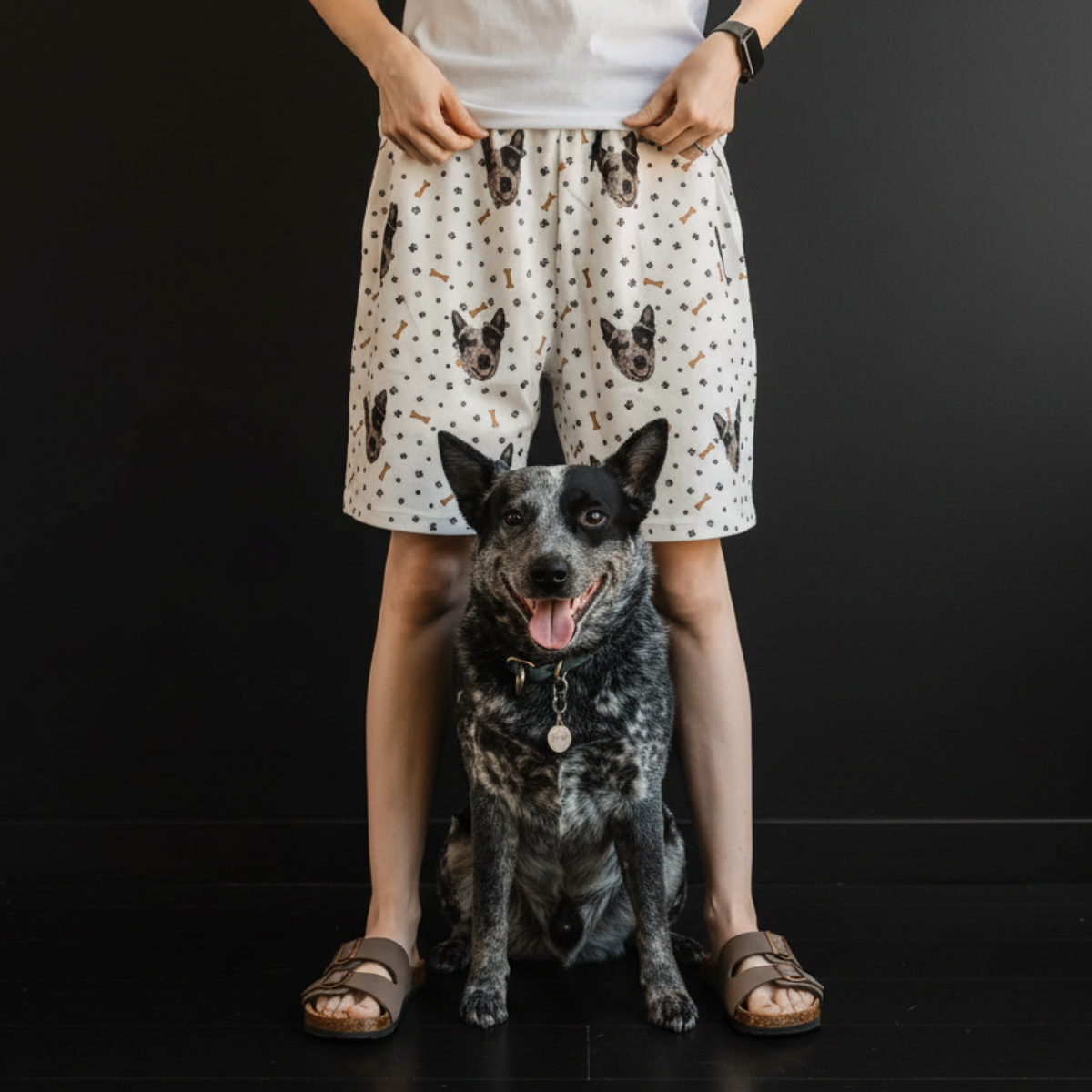 Person wearing shorts with dog pattern standing next to a dog on a black background