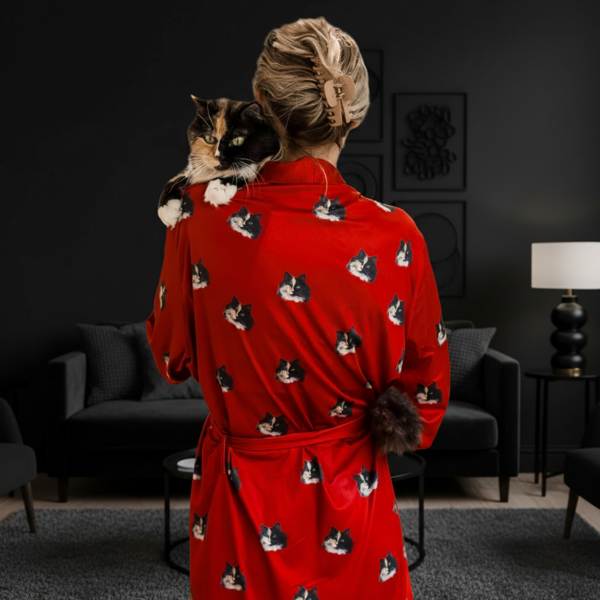 Person wearing a red robe with cat patterns, holding a cat, in a dark living room.