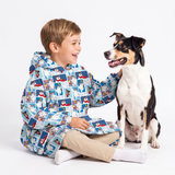 A young boy wearing a blue hooded raincoat with a cartoon dog print sits on the floor next to a black and white dog, smiling and petting it, against a plain white studio background.