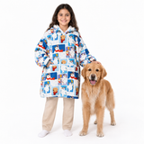 A young girl stands next to a golden retriever dog wearing a hooded blanket coat covered in a blue and white cartoon dog print, paired with beige pants and white socks, on a solid white background.