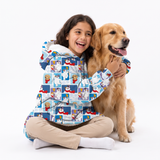 A young girl with dark hair sits cross-legged on a white studio background, hugging a golden retriever dog. She is wearing a light blue hooded jacket covered in a colorful cartoon dog and snowboard pattern, paired with beige pants and white socks.