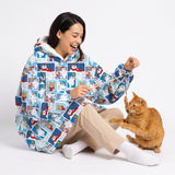 A woman sitting on a white studio floor wearing a light blue hooded sweatshirt covered in a colorful cat-themed patchwork print, interacting playfully with an orange tabby cat.